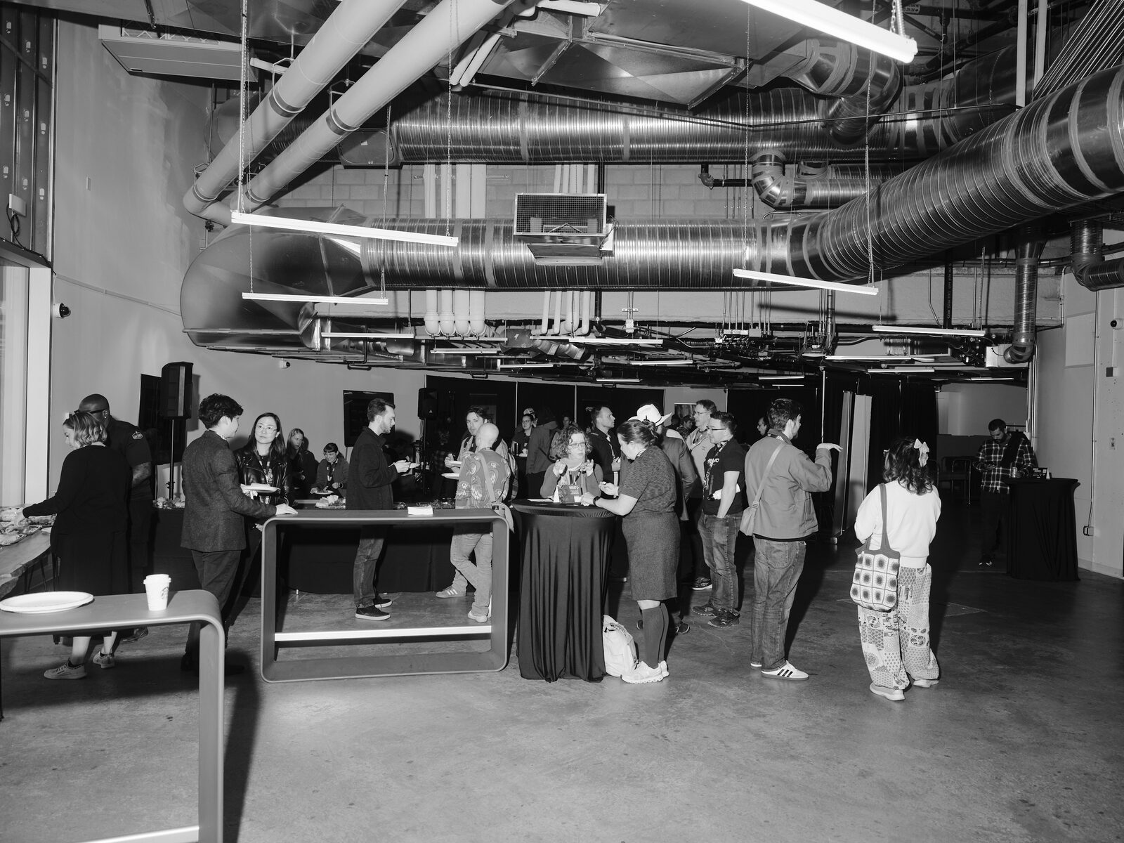 Attendees mingle around high-top tables and grab food from a buffet in an industrial room lined with silver ductwork and hanging lights.