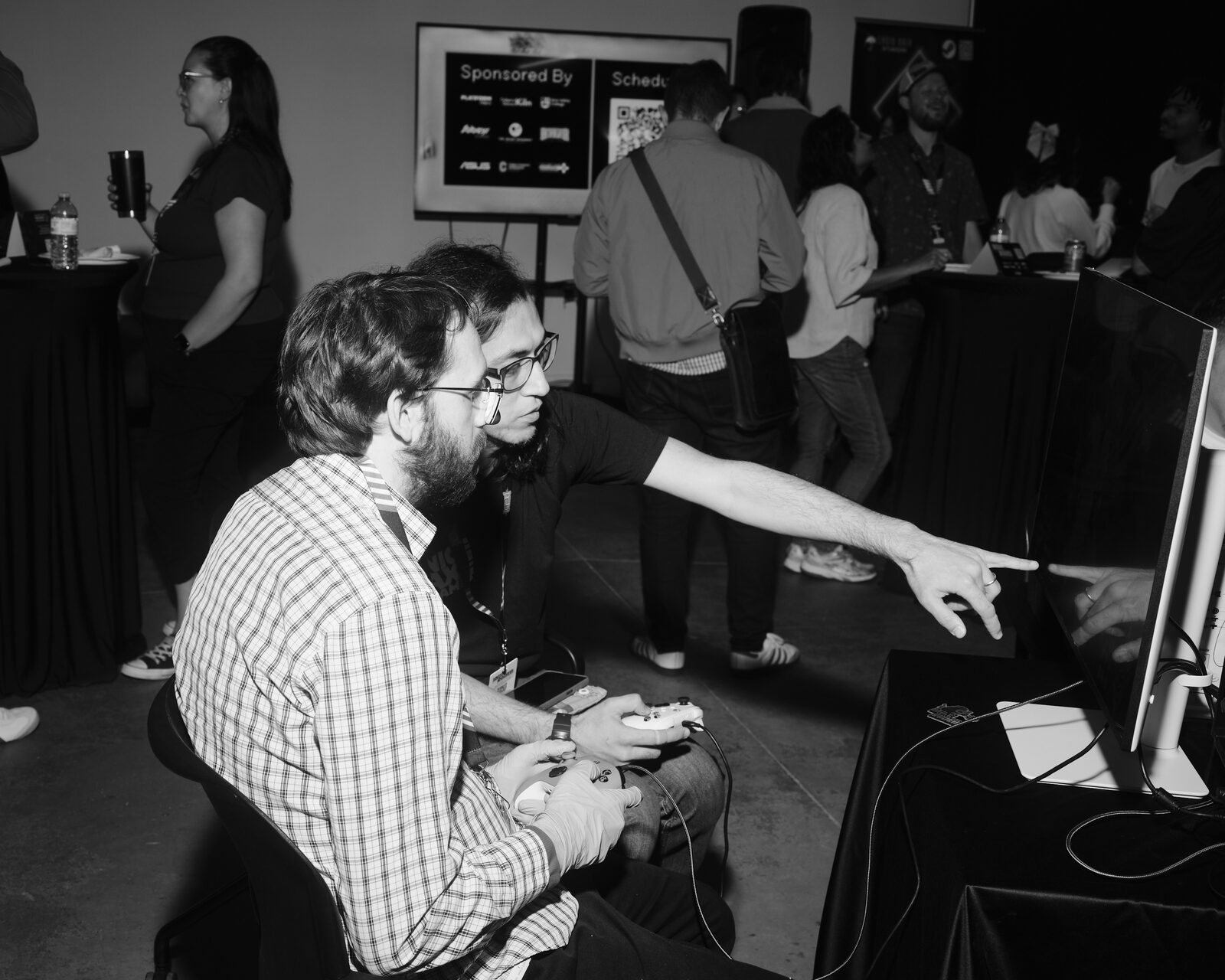 Two players in glasses sit side by side with controllers as one points at the demo screen, a "Sponsored By" board and the busy showcase floor behind them.