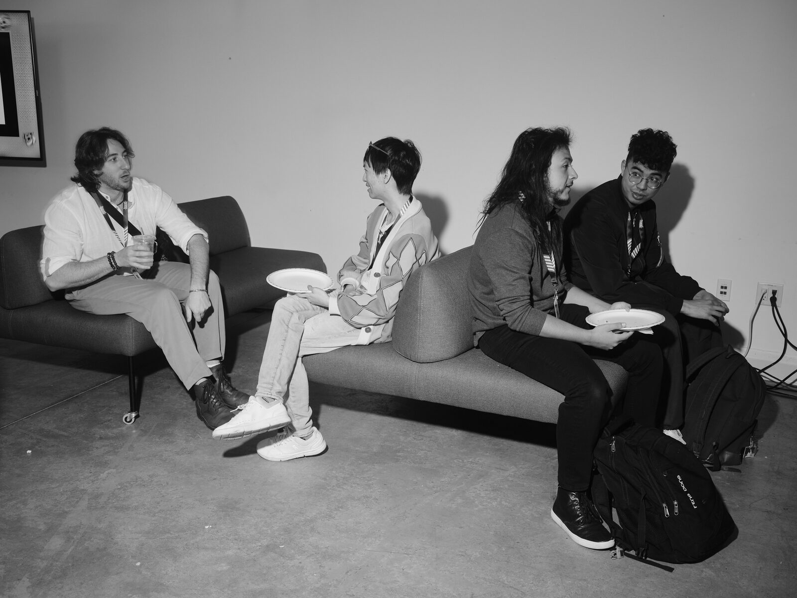 Four attendees with lanyards sit on low grey couches against a white wall, chatting over paper plates and drinks.