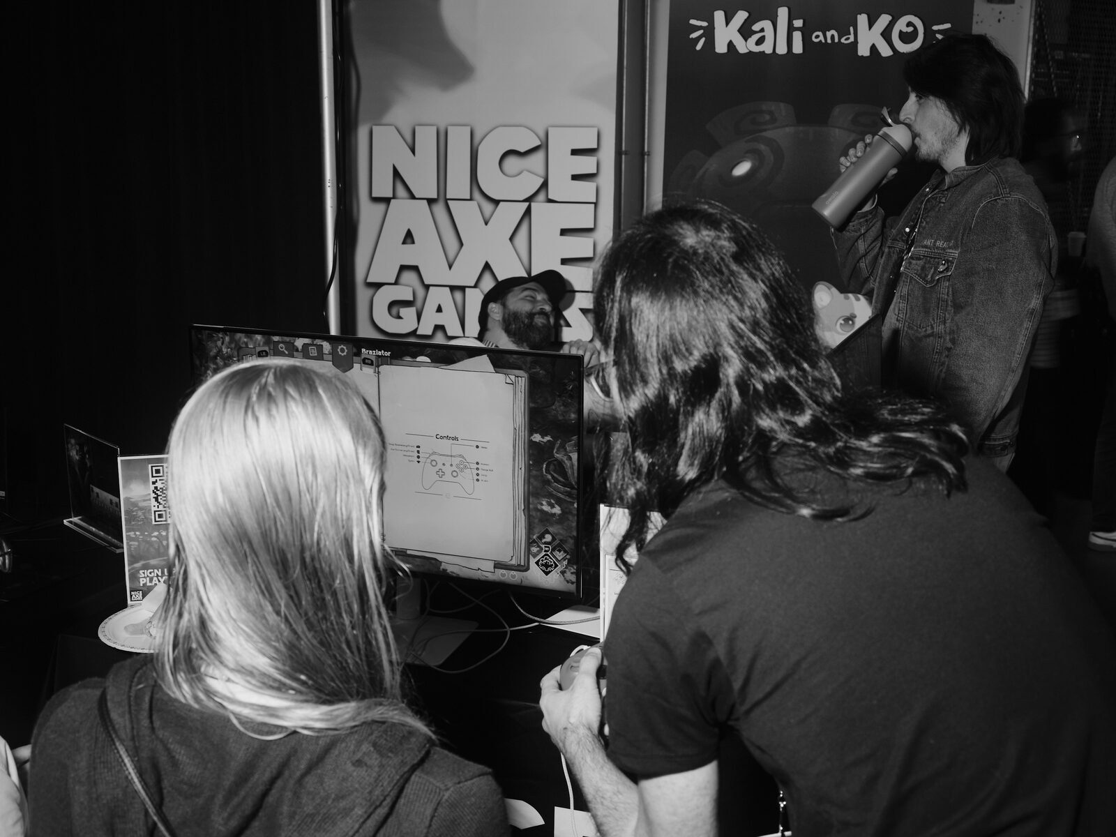 Two long-haired players try a demo at a monitor showing a controls screen, with Nice Axe Games and Kali and KO banners behind them and an onlooker sipping from a bottle.