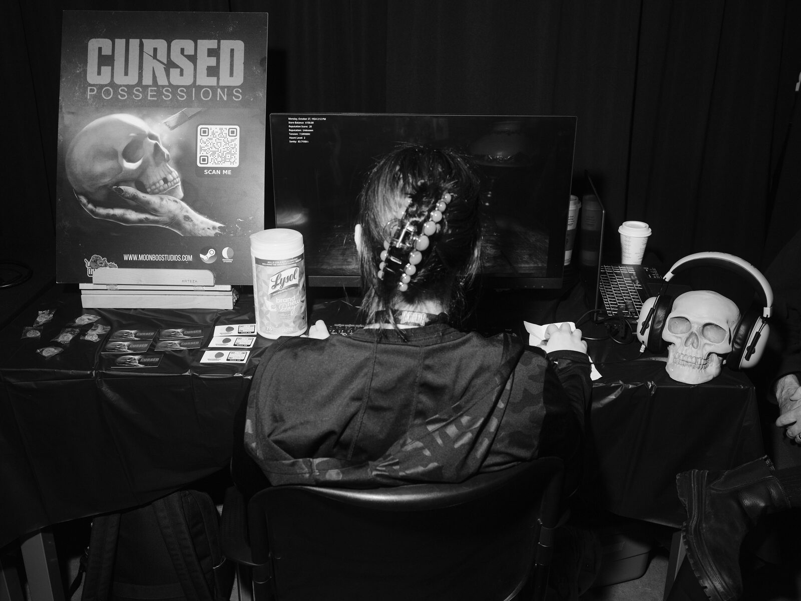 A player with a beaded hair clip sits at the Cursed Possessions booth, the skull poster and QR code on one side, a plastic skull wearing headphones on the other.