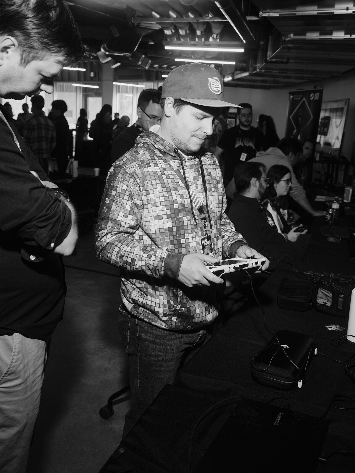 An attendee in a pixel-pattern hoodie grins down at a handheld gaming PC at a long demo table, other players seated along it.