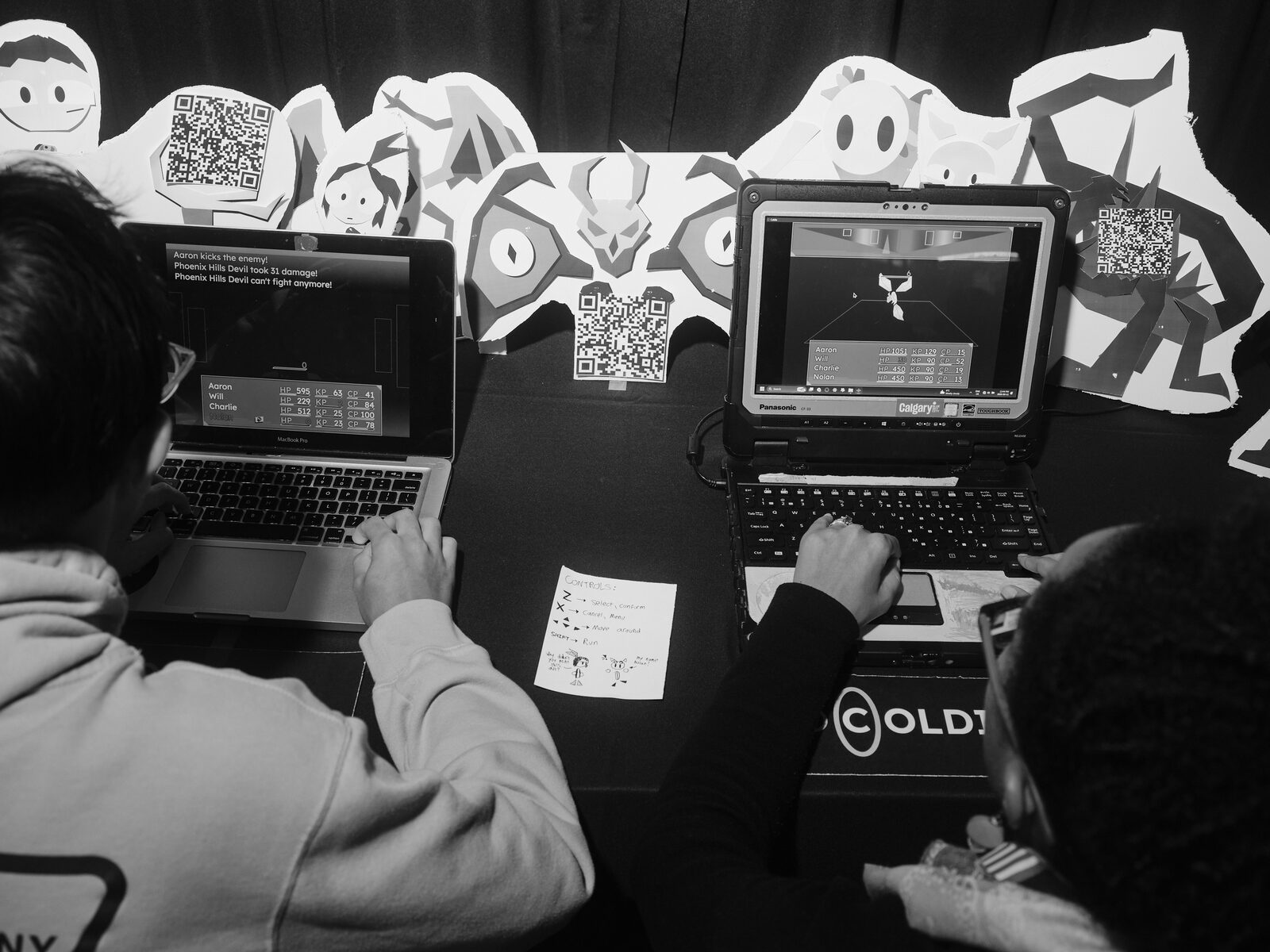 Two players at laptops play a turn-based battle game reading "Phoenix Hills Devil can't fight anymore!", paper cutout monster art with QR codes lining the table behind.