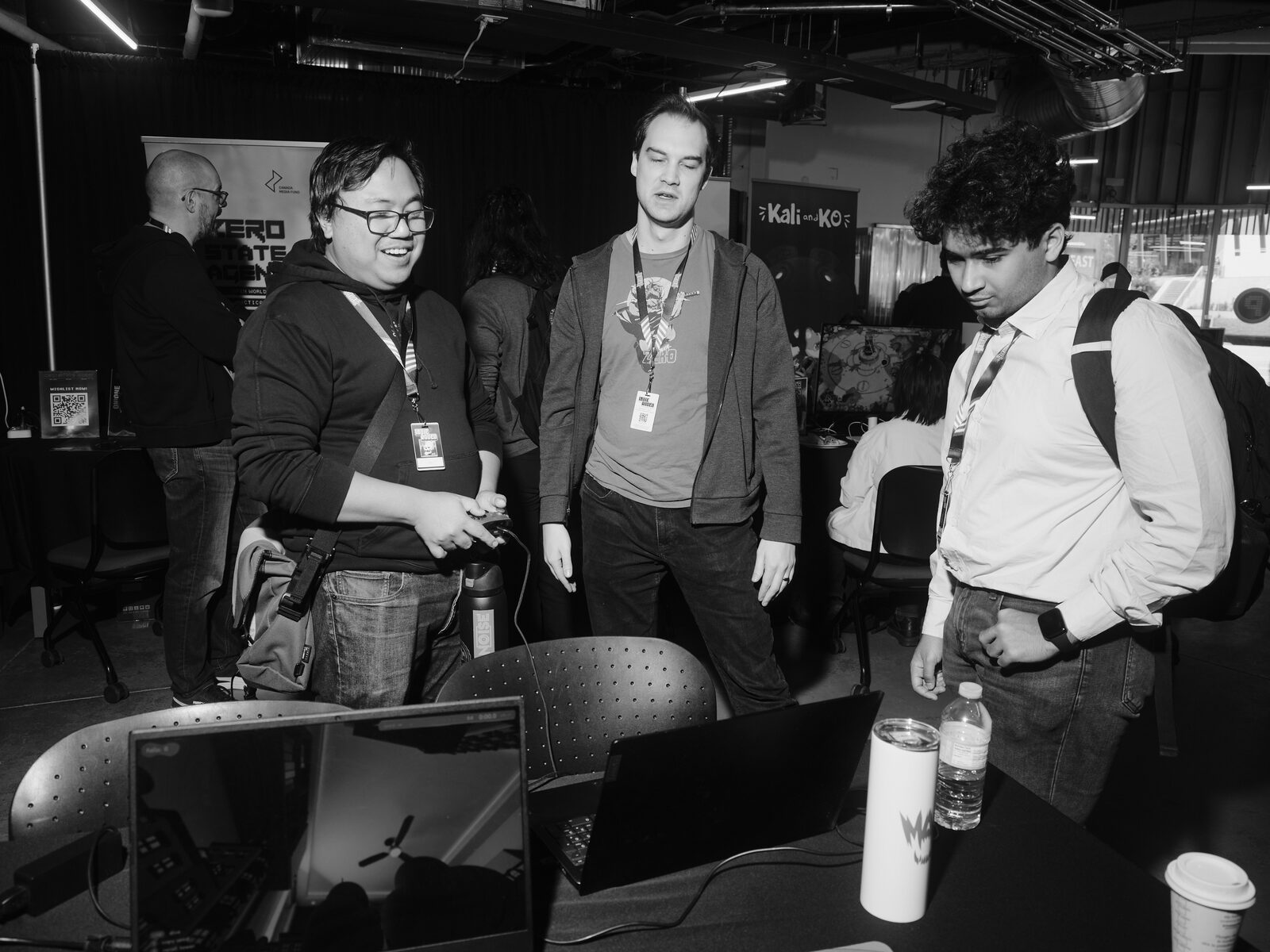 Three attendees with lanyards laugh and watch as one tries a controller at a laptop demo, Zero State Agent and Kali and KO banners behind them.