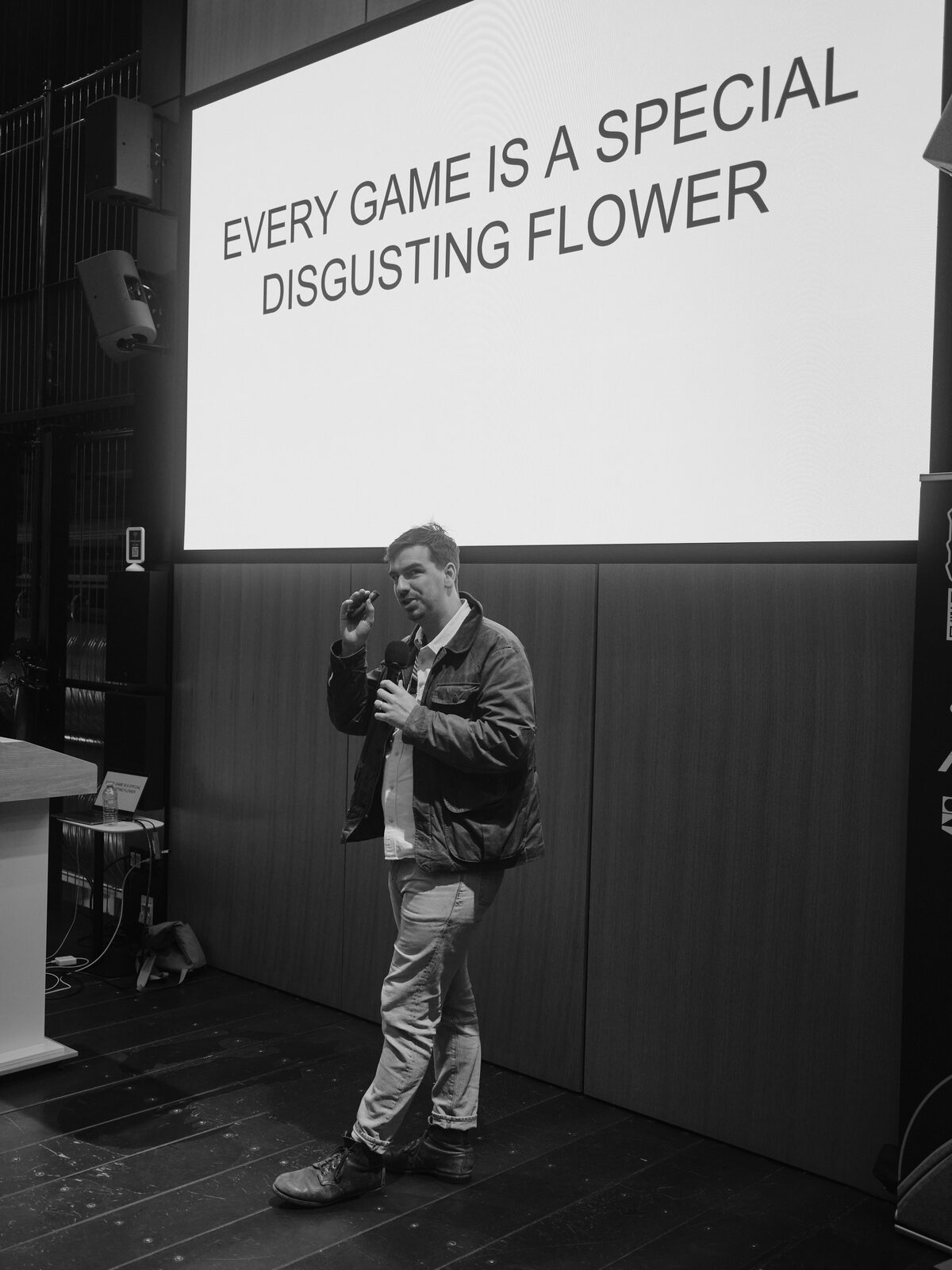 A speaker in a waxed jacket gestures with a clicker below a slide reading "EVERY GAME IS A SPECIAL DISGUSTING FLOWER".