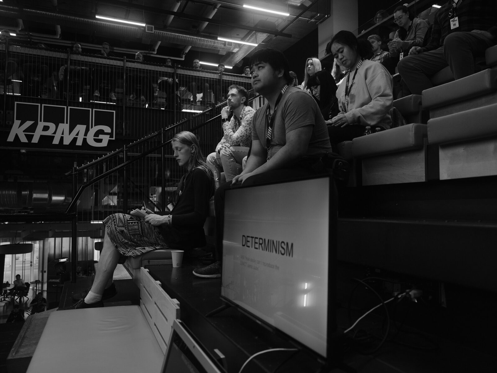 Attendees on the bleachers watch a talk, one reading from a notepad, a stage monitor in the foreground showing a slide titled "DETERMINISM" and the KPMG sign behind.