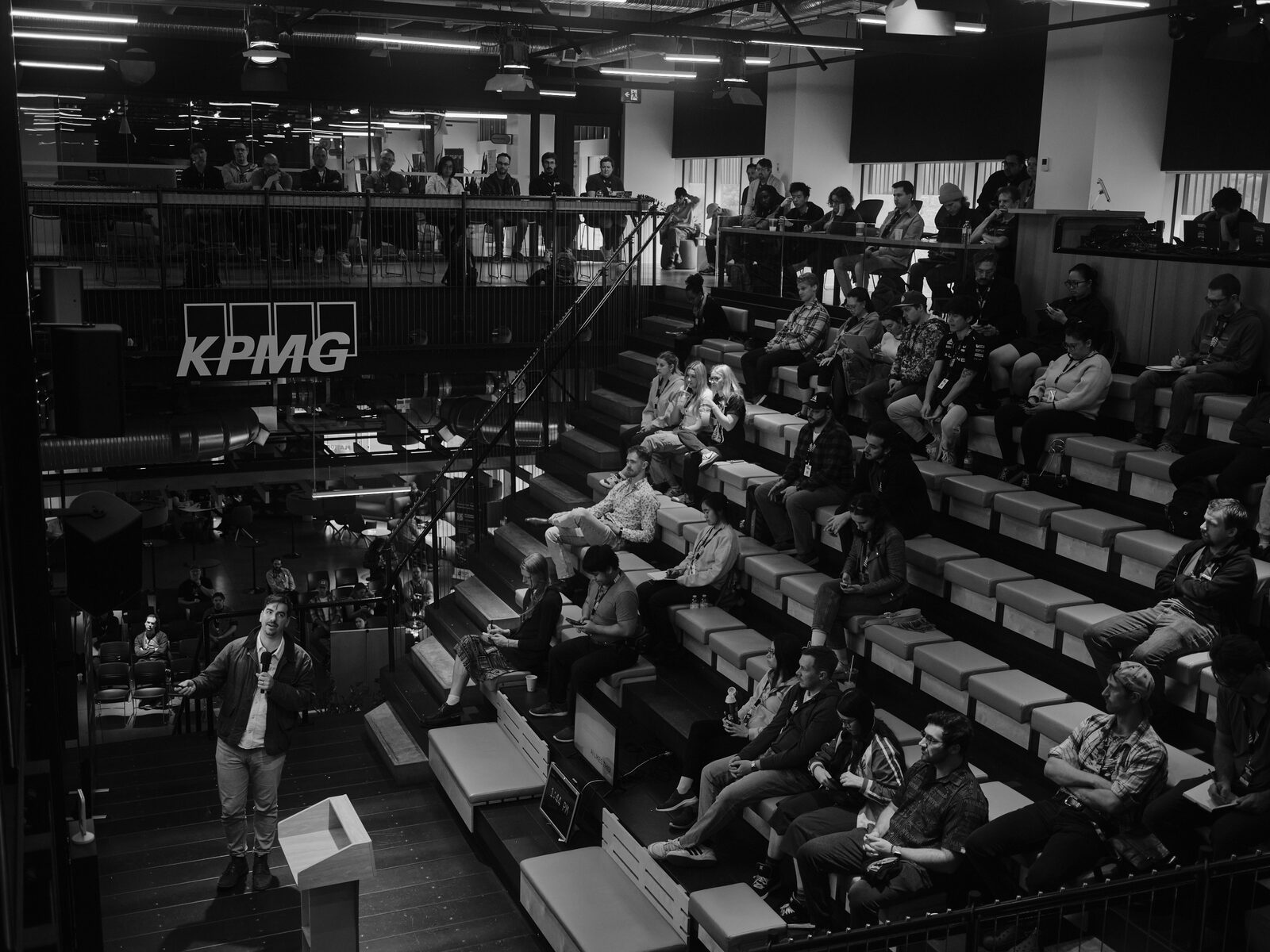 High view of the full atrium: a speaker in a jacket talks from the stage floor as attendees fill the bleachers and the balcony rail beneath the KPMG sign.