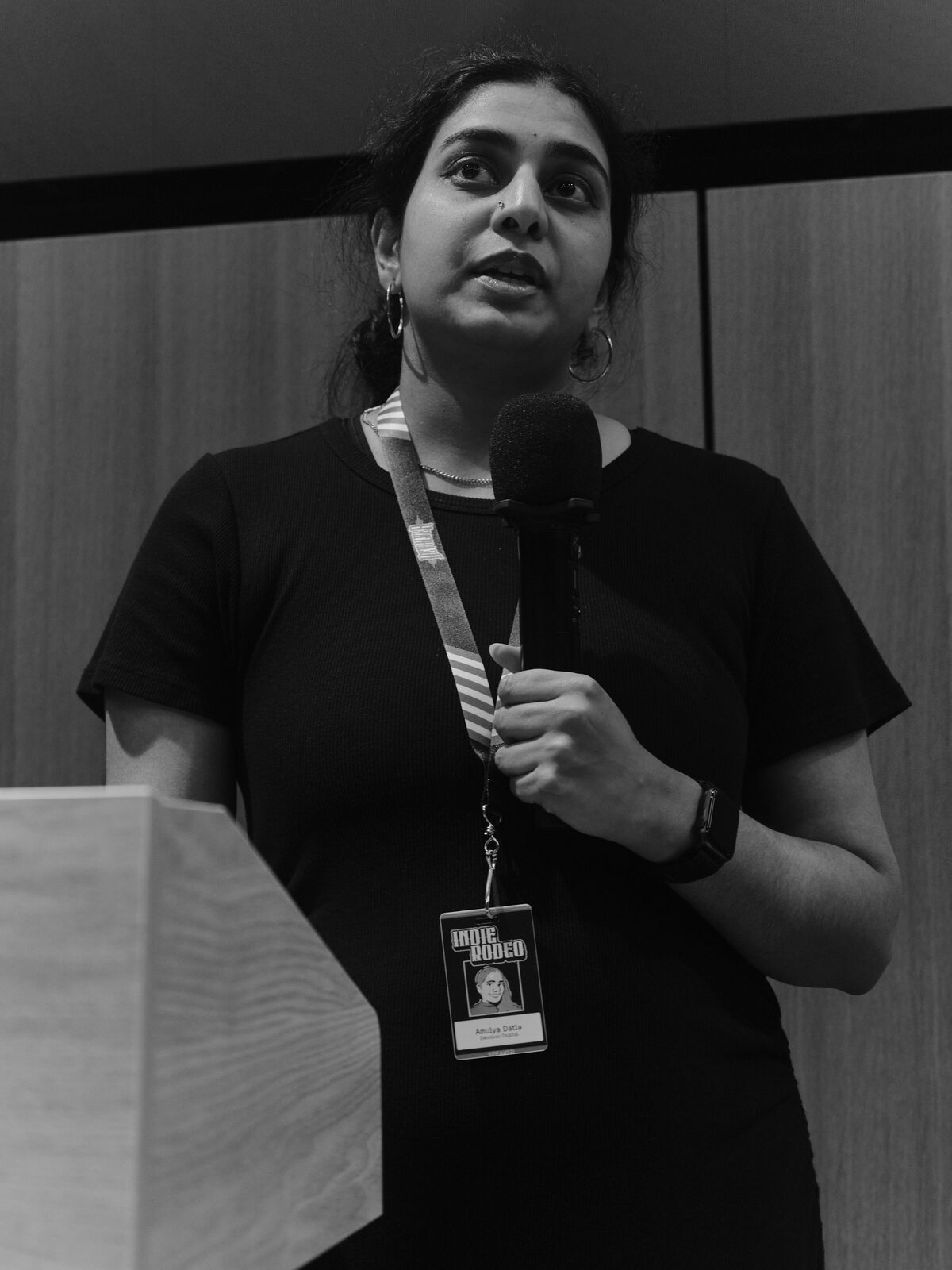 A speaker in a black tee with hoop earrings speaks into a handheld mic beside the podium, their Indie Rodeo badge hanging from a striped lanyard.