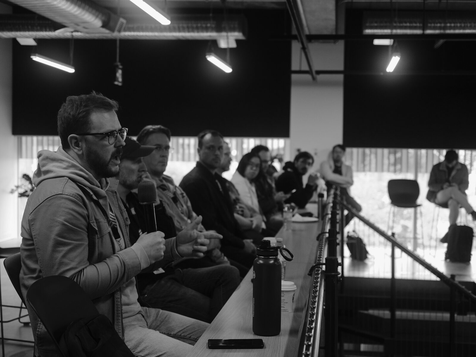 A row of panelists sits along a high counter by the stair rail, one in glasses and a hoodie mid-answer on the mic, others listening.