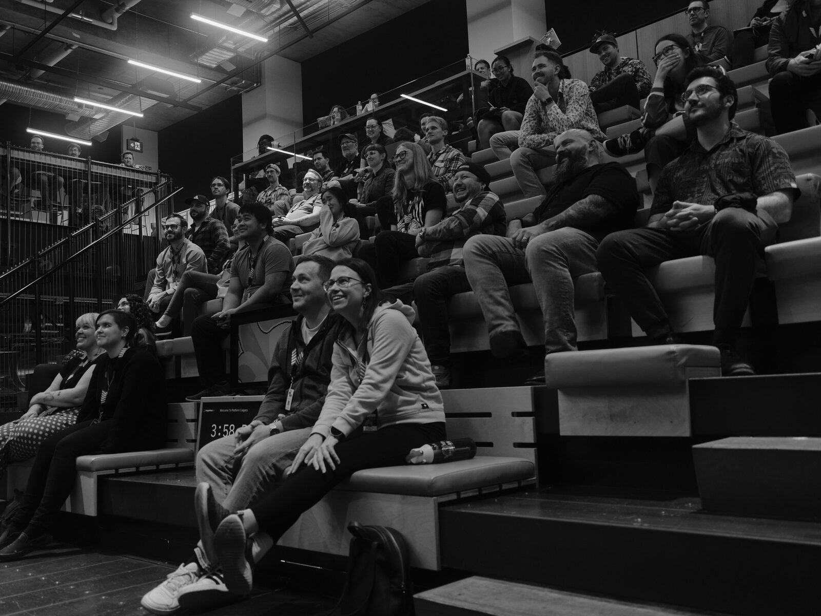Attendees on the bleachers grin and laugh during a talk, a stage monitor in front of them showing a countdown timer.