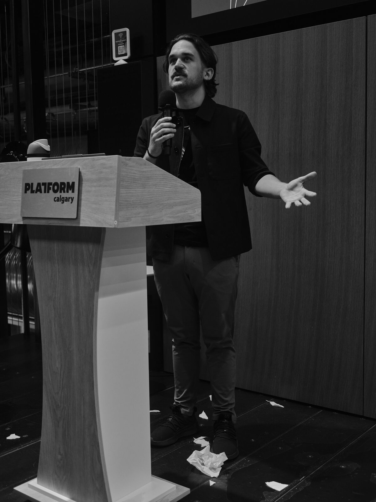 A speaker in a dark overshirt spreads one hand and looks upward beside the Platform Calgary podium, crumpled paper scattered across the stage floor.