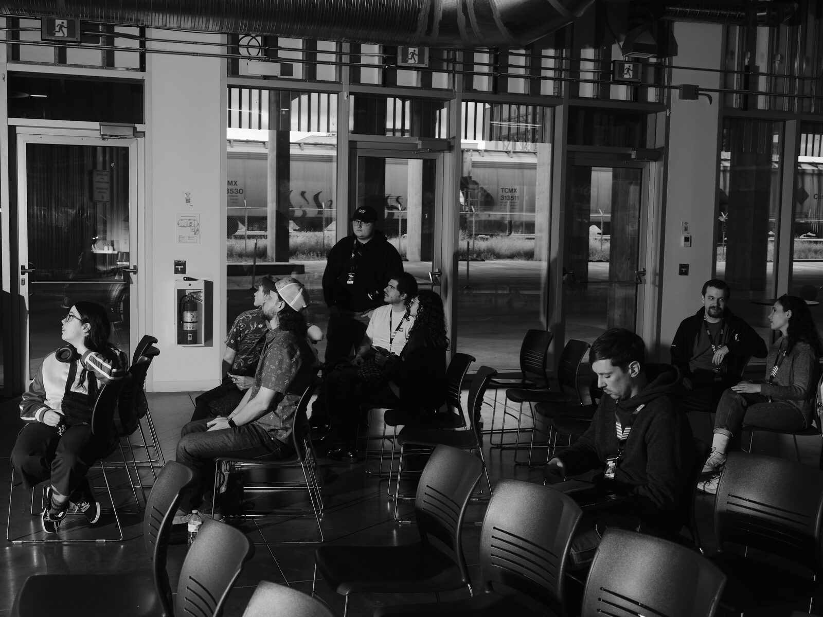 Attendees sit scattered among rows of chairs in a sunlit ground-floor room, freight train cars visible through the tall windows behind them.
