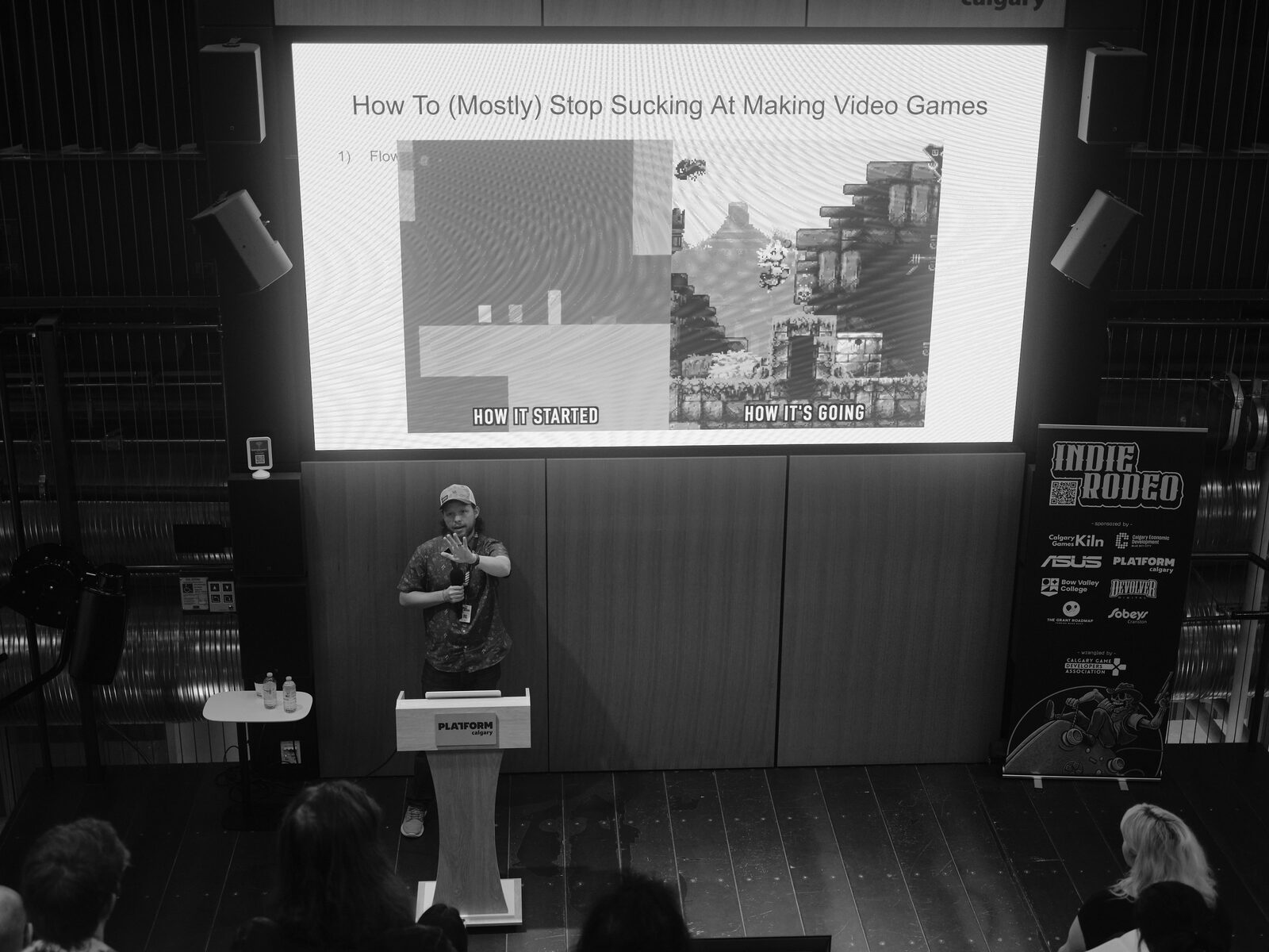From the balcony, a speaker in a ball cap stands by the Platform Calgary podium under a slide reading "How To (Mostly) Stop Sucking At Making Video Games" and "HOW IT STARTED".