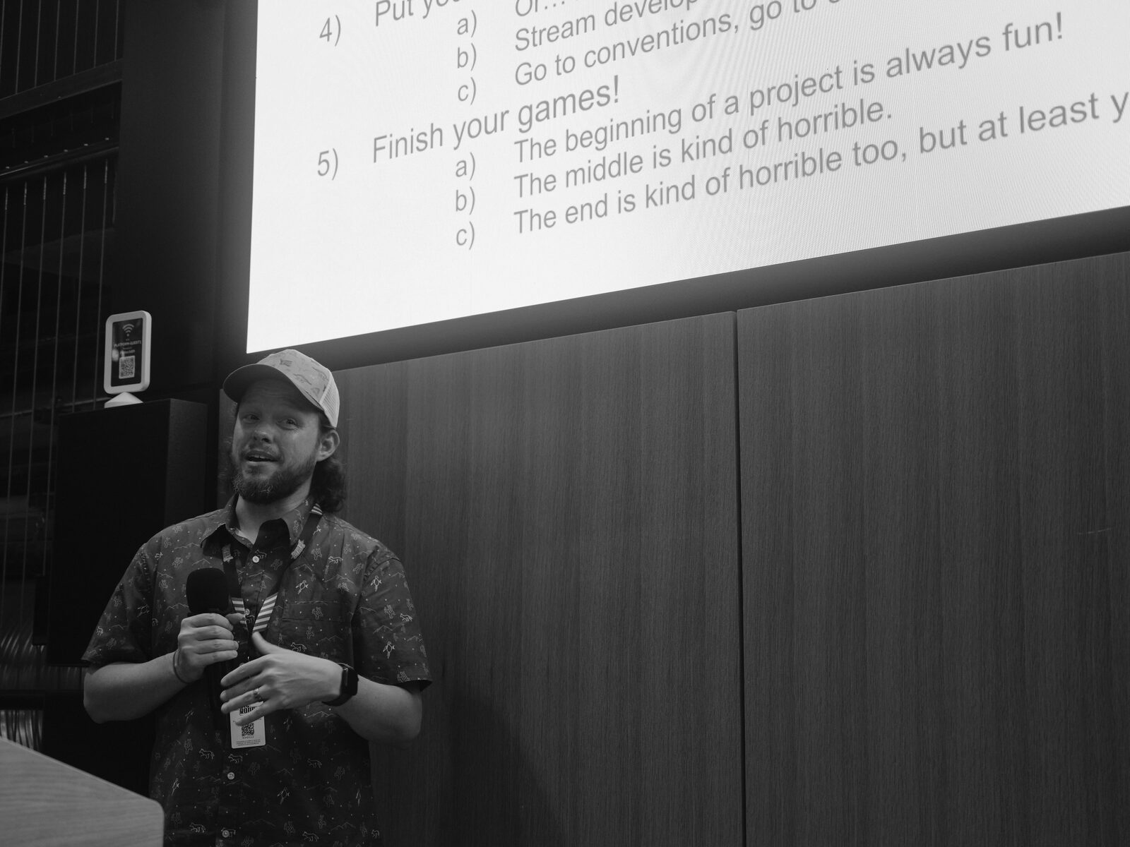 A speaker in a ball cap and patterned short-sleeve shirt grins mid-talk, mic in hand, beneath a slide reading "Finish your games!" and "The beginning of a project is always fun!".