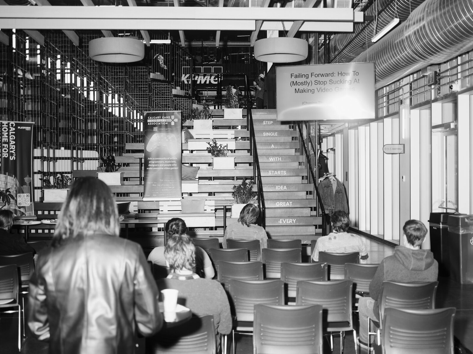 An attendee in a leather jacket stands behind rows of overflow seating facing a screen that reads "Failing Forward", the lettered staircase and a Calgary Game Developers Association banner beside it.