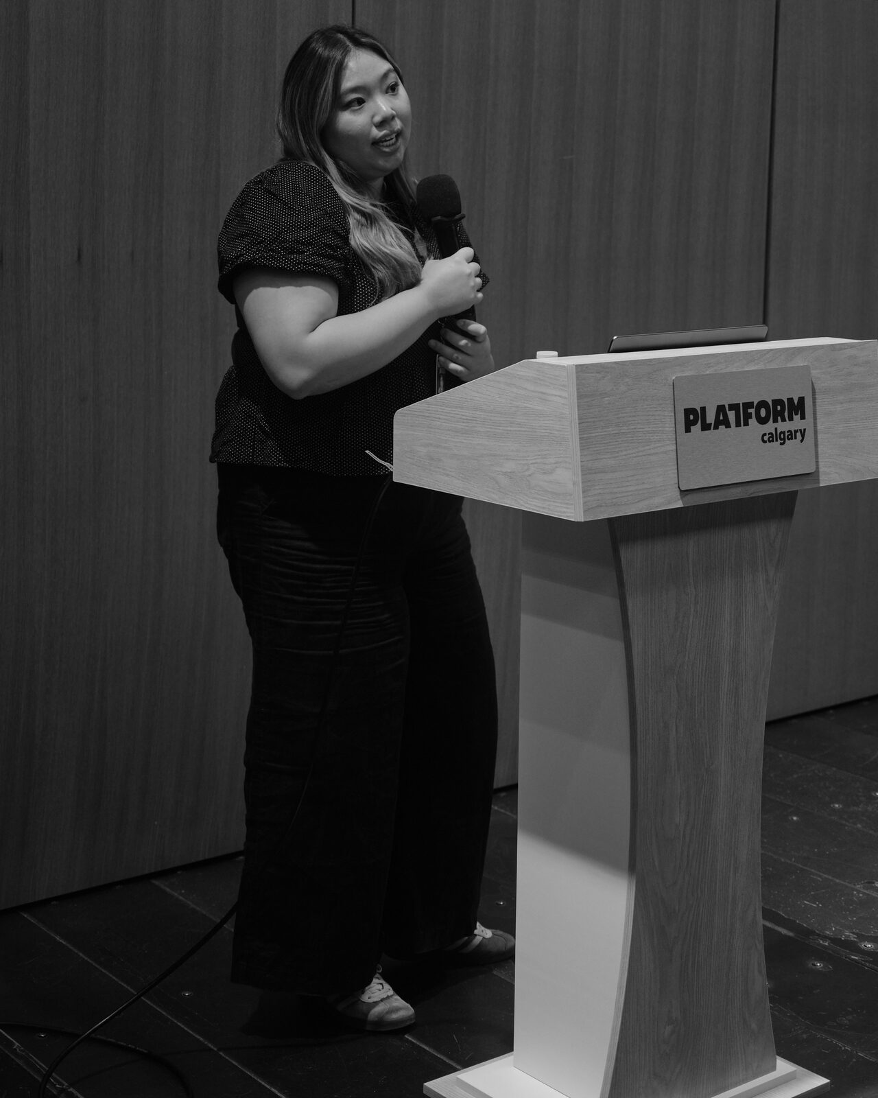 A speaker in a polka-dot top holds a mic beside the Platform Calgary podium, mid-sentence, lit against the dark wood-panel wall.