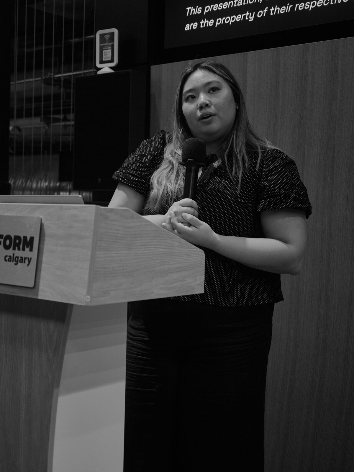 A speaker in a polka-dot top stands at the Platform Calgary podium with a mic, looking out at the room.