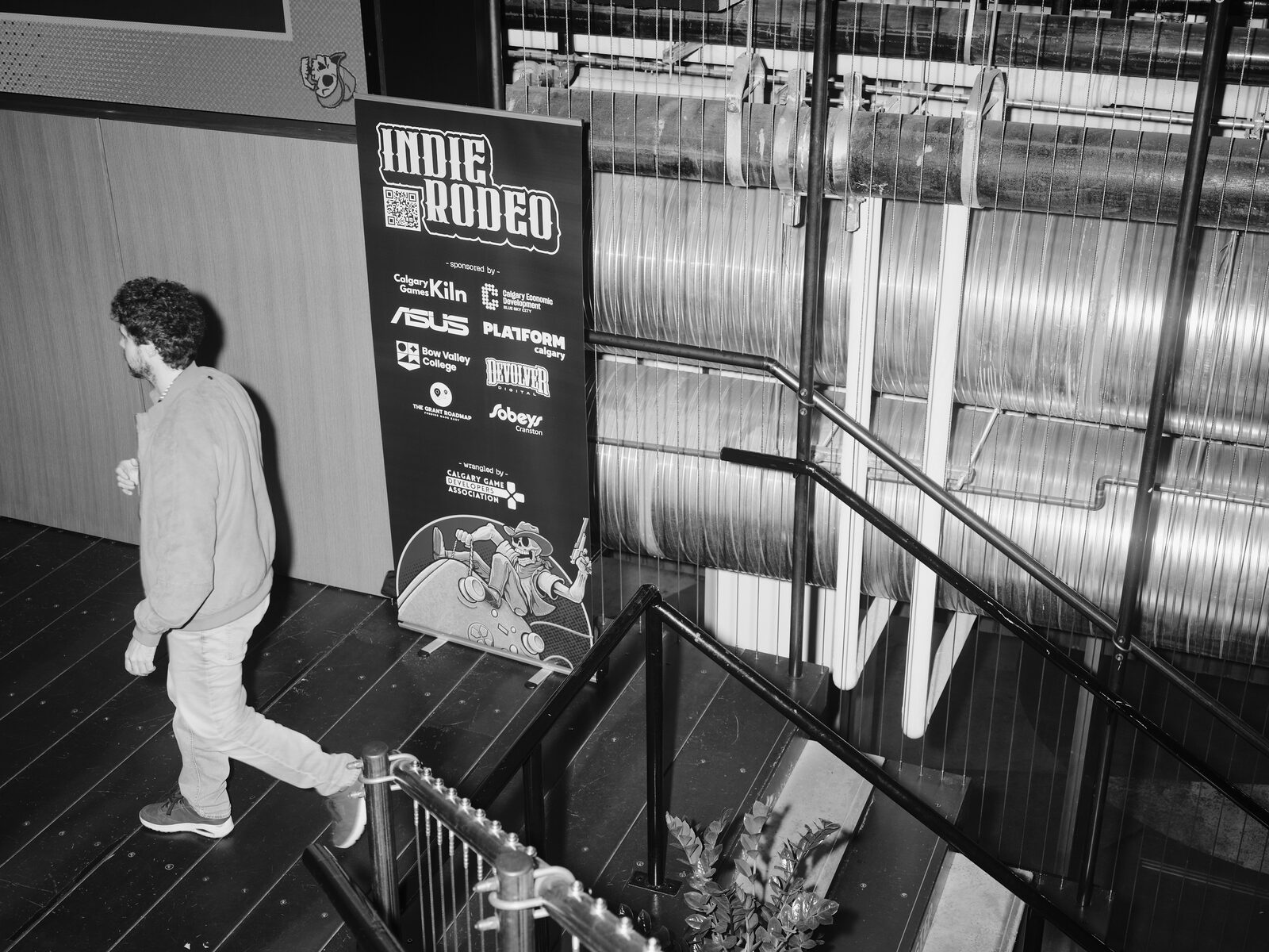 Looking down the stairs as an attendee walks past the Indie Rodeo sponsor banner, its undead-cowboy mascot and logos for Calgary Games Kiln, ASUS, Platform Calgary, and Devolver Digital.