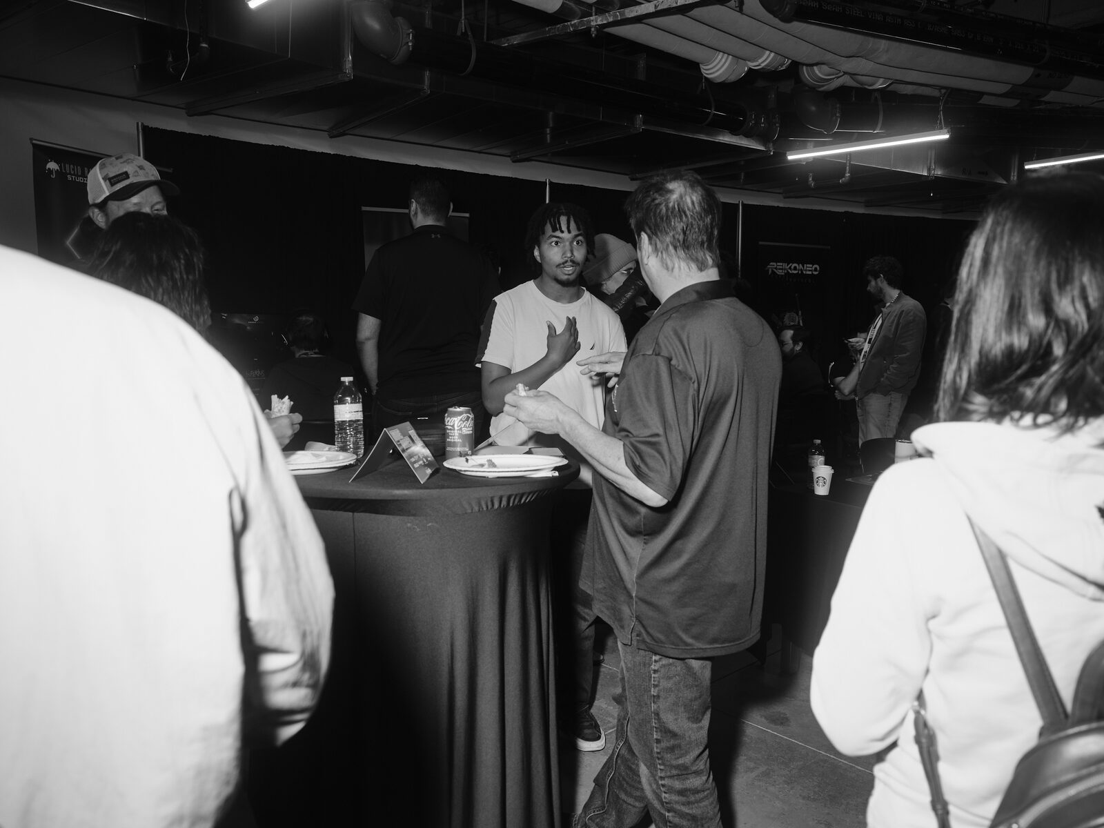 Attendees chat over paper plates and cans around a draped cocktail table on the showcase floor, booth banners in the dim background.