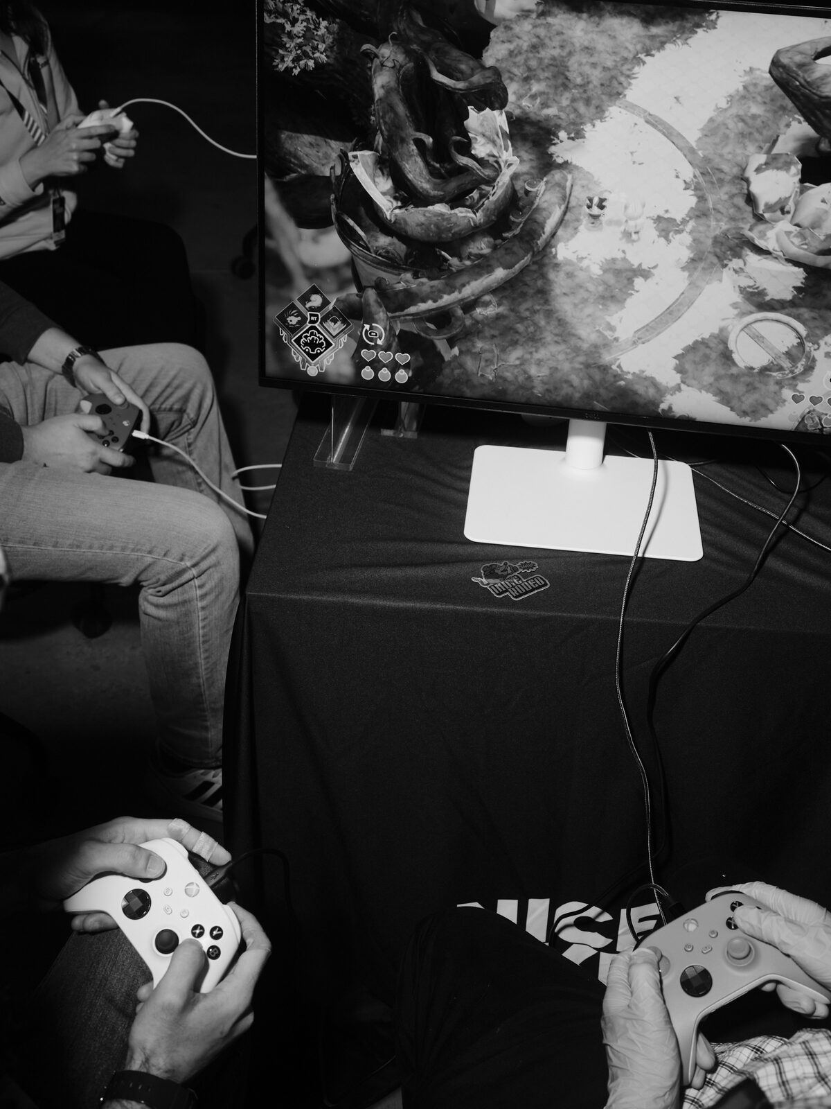 Overhead view of four hands on controllers around a monitor showing a top-down game with a hearts health bar, an Indie Rodeo sticker on the black tablecloth.