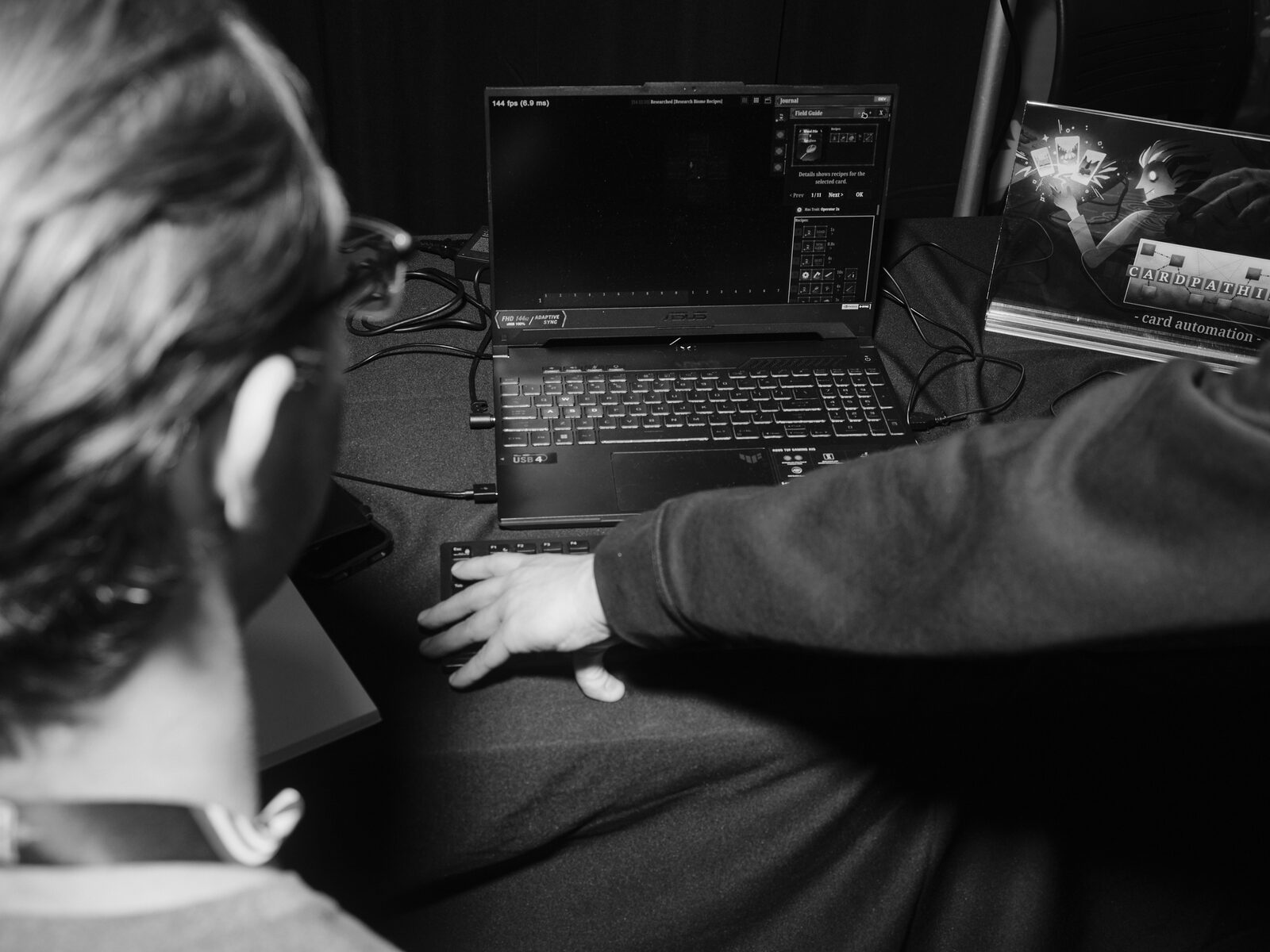 Over the shoulder of a player at a laptop demo, a card game's "Field Guide" menu on screen and a Cardpathic "card automation" sign propped beside it.