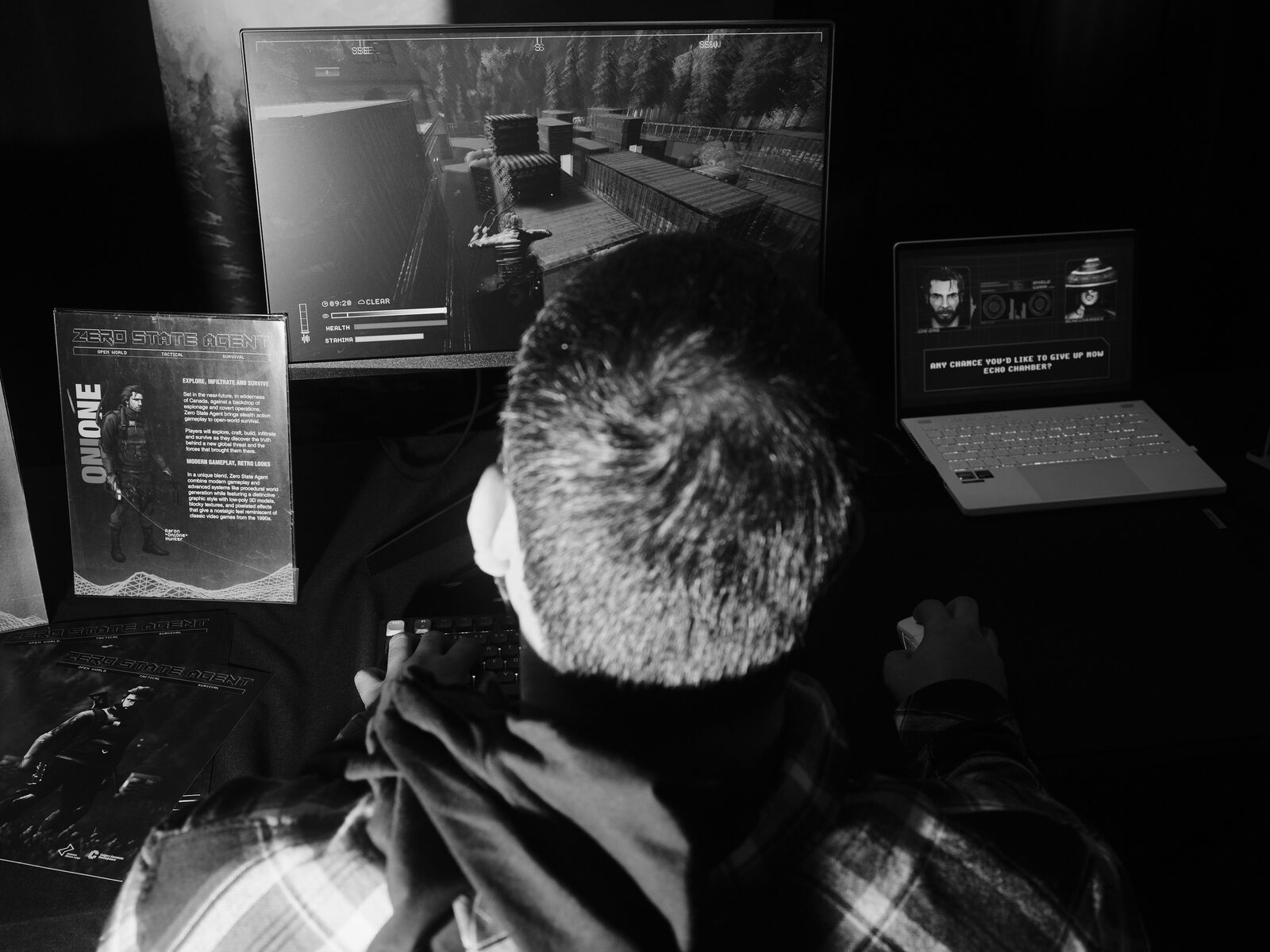 From behind a player at the Zero State Agent booth, a third-person shooter on the monitor, the game's info card to one side and a dialogue screen on a laptop.
