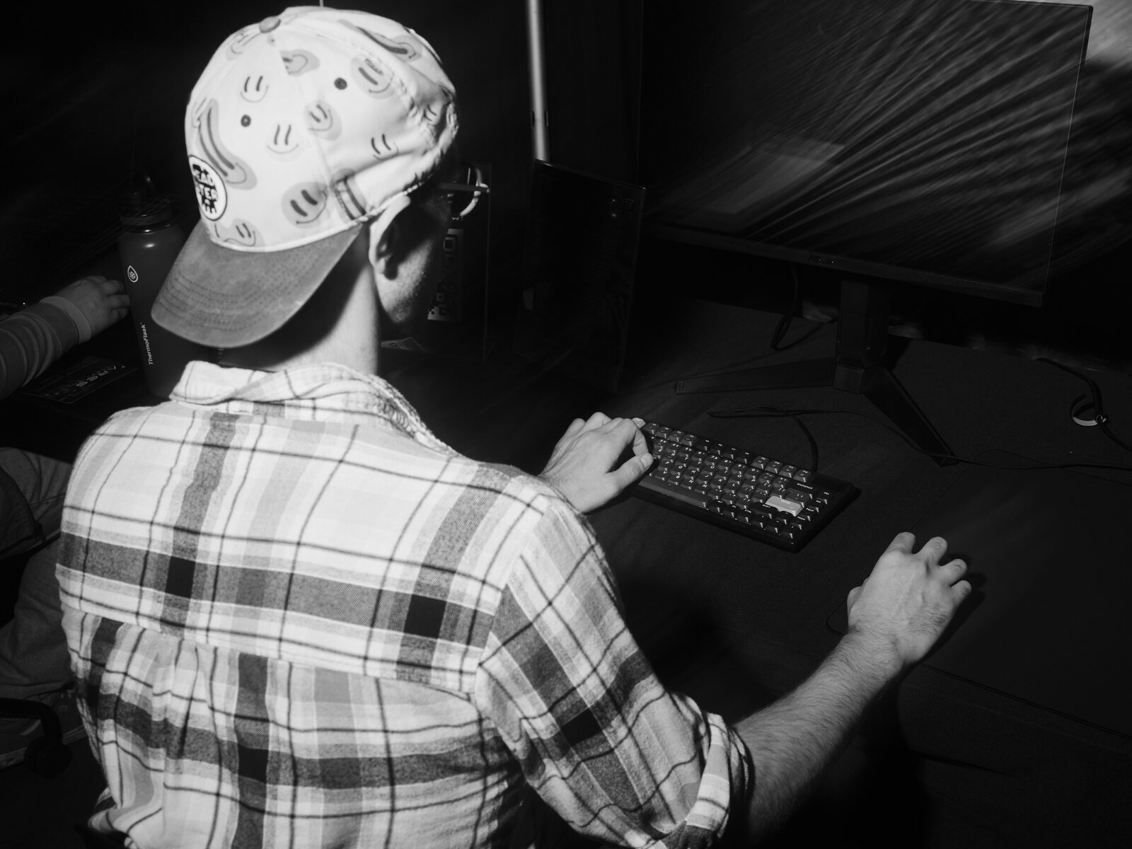 Over the shoulder of a player in a smiley-face cap and plaid shirt, hands on a mechanical keyboard and mouse at a darkened demo station.
