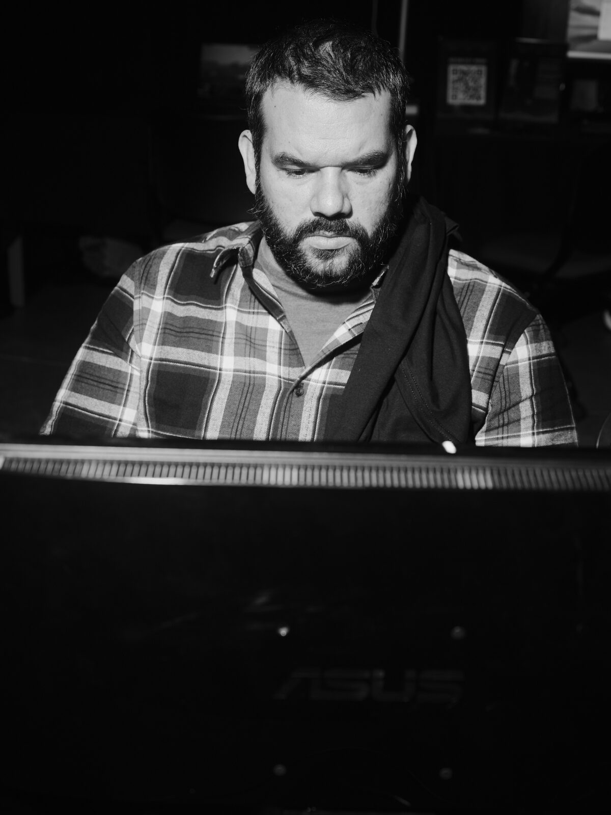 A bearded attendee in a plaid shirt looks down, lit from below by the ASUS monitor they're playing on, the room dark behind them.