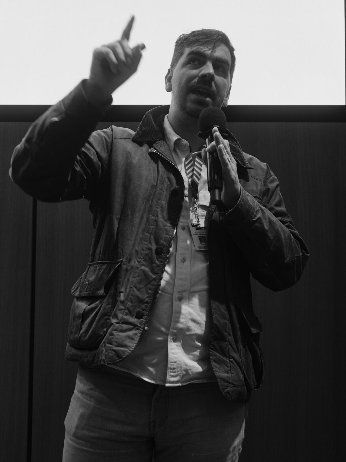 Low-angle close shot of a speaker in a waxed jacket raising one finger to make a point, mic in hand, Indie Rodeo badge on their lanyard.