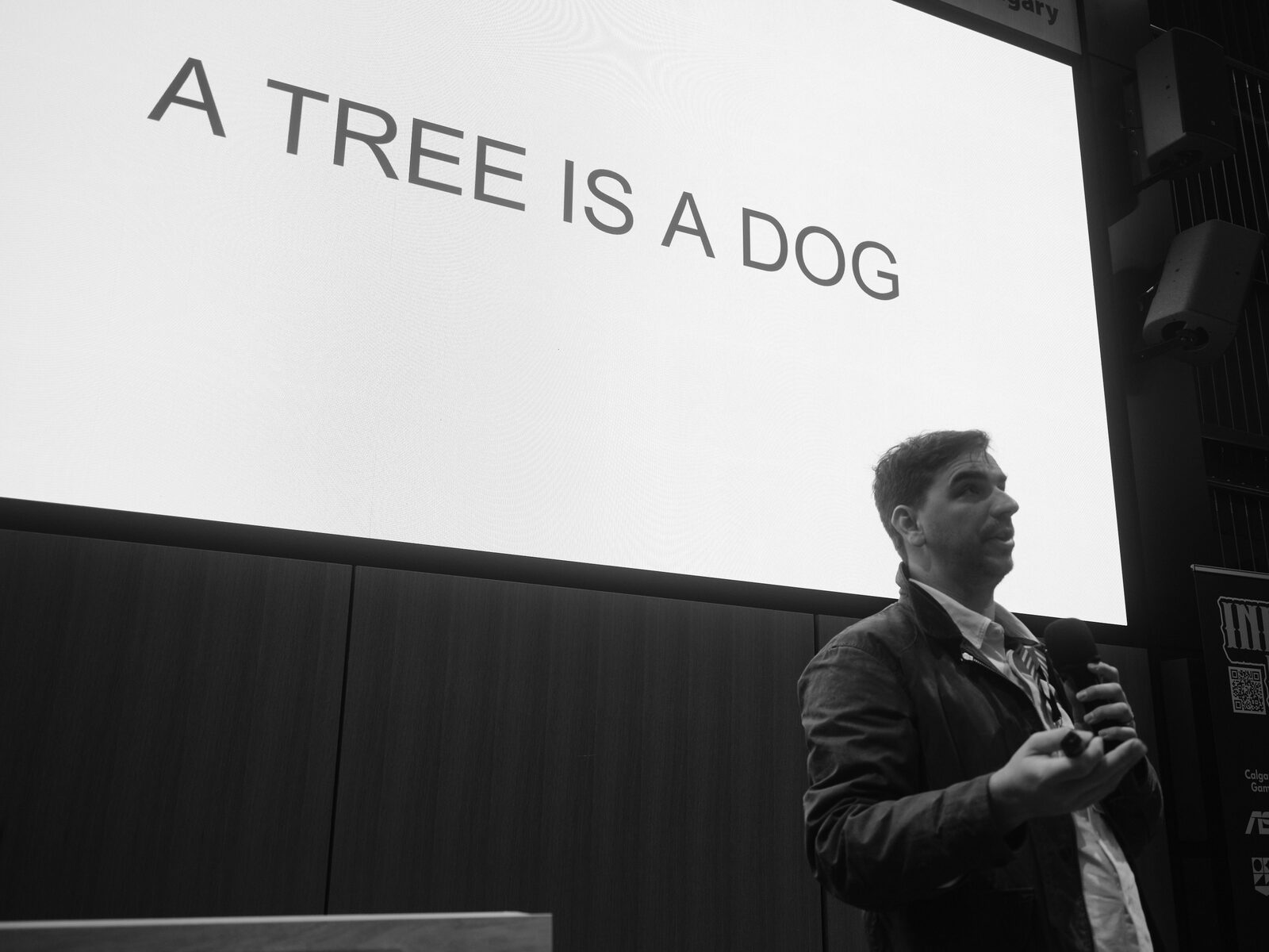 A speaker in a waxed jacket looks up toward a stage screen reading "A TREE IS A DOG", mic and clicker in hand.