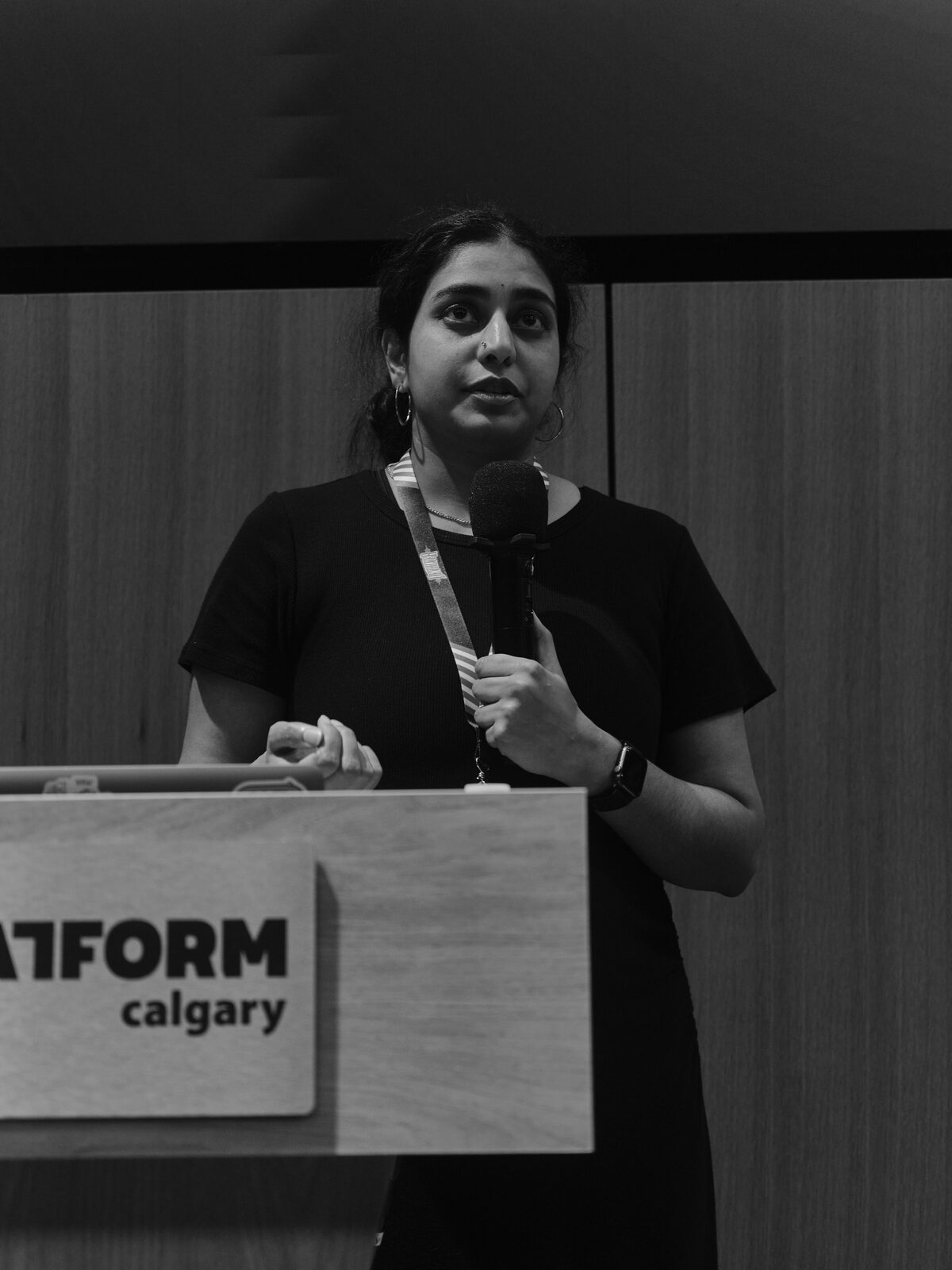 A speaker in a black tee and lanyard talks into a mic behind the Platform Calgary podium, one hand raised mid-point.