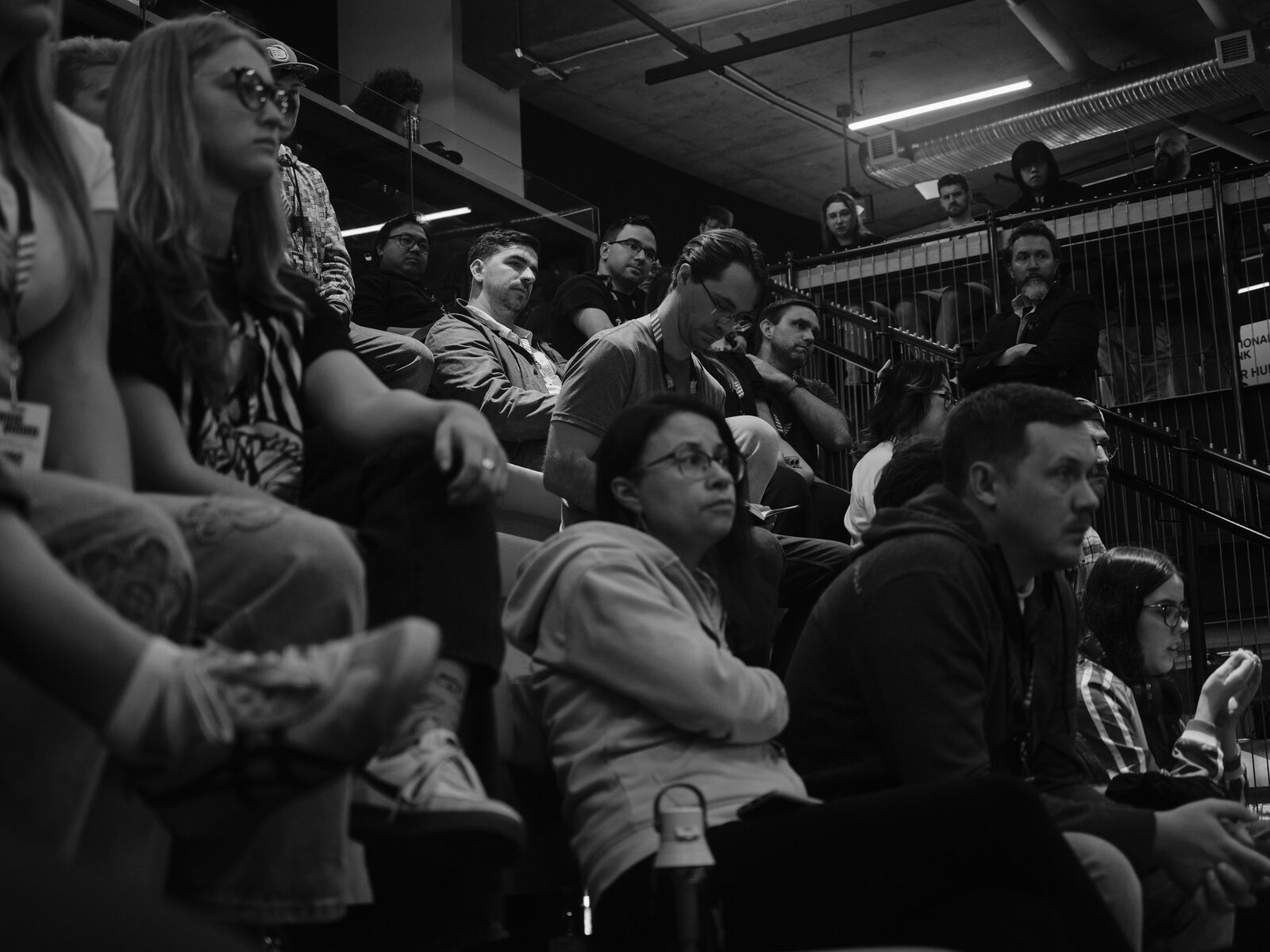 Attendees on the bleachers listen closely to a talk, one with legs crossed in the foreground, the balcony rail above.