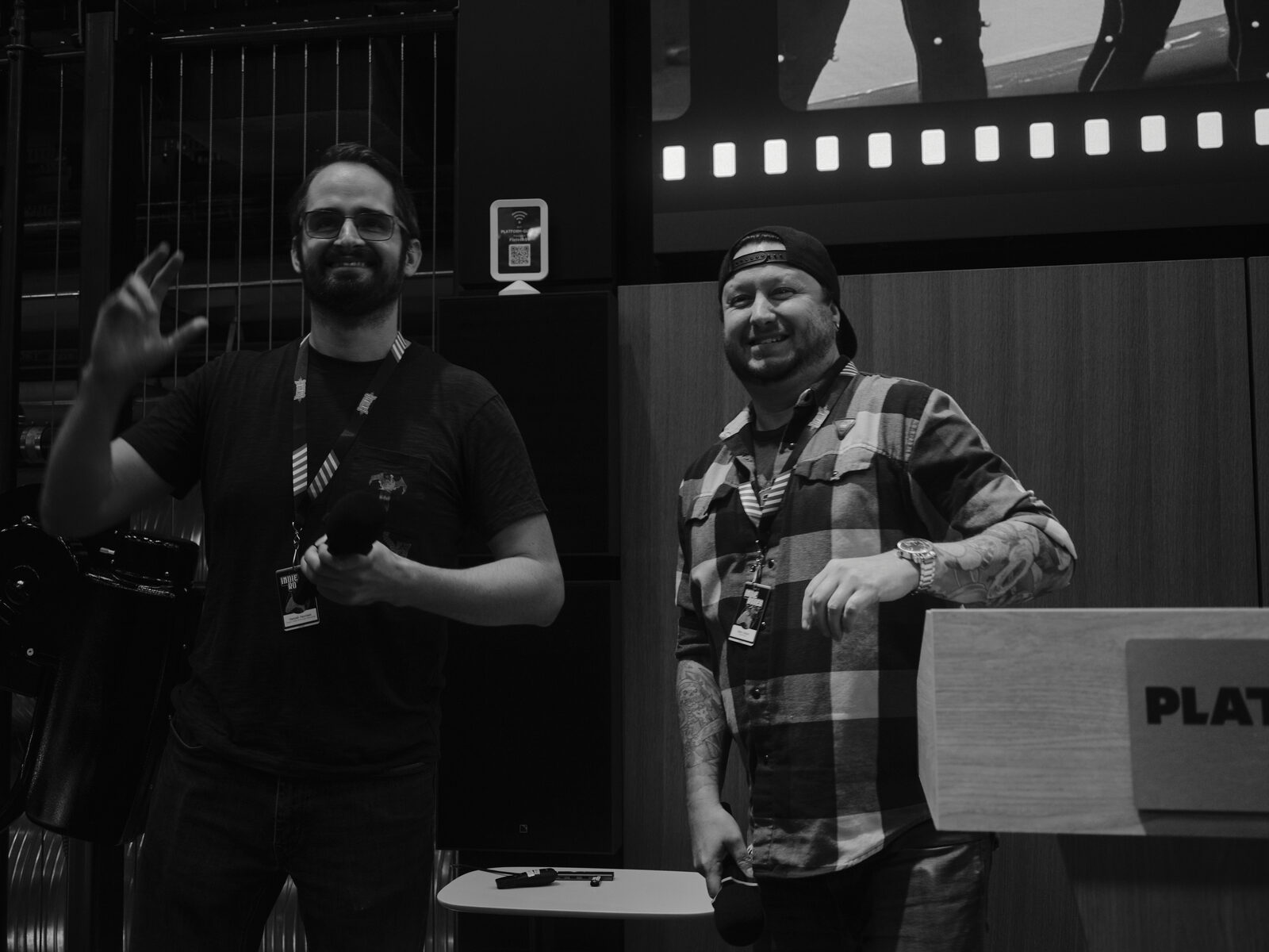Two speakers grin beside the Platform Calgary podium, one in a black tee waving, the other in a plaid shirt and backwards cap.
