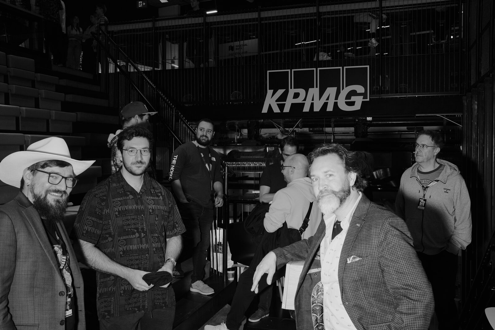 Attendees gather at the foot of the bleachers under the KPMG sign, one in a white cowboy hat up front and another in a tweed blazer leaning on the rail.