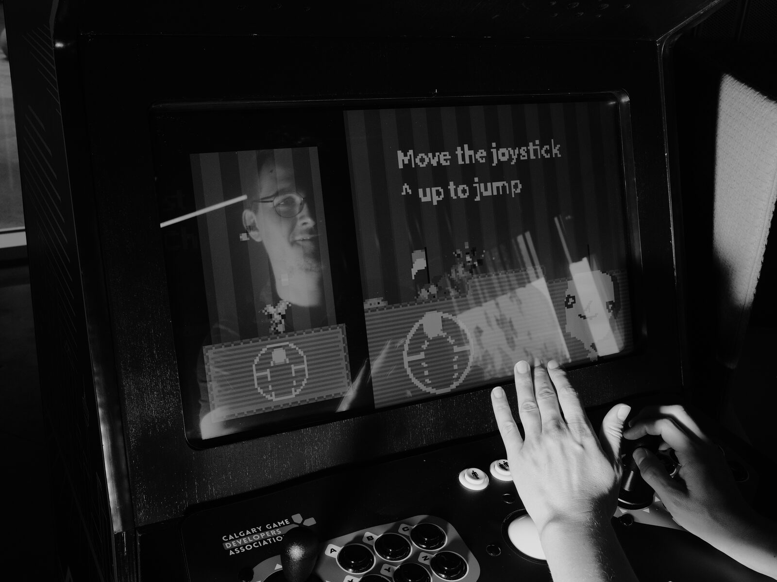 Close on hands at a Calgary Game Developers Association arcade cabinet, the pixel screen reading "Move the joystick up to jump", a face reflected in the glass.