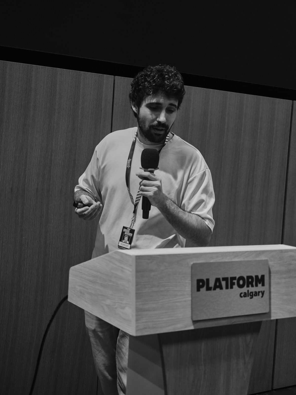 A curly-haired speaker glances down at their clicker at the Platform Calgary podium, mic in hand and Indie Rodeo badge on their lanyard.