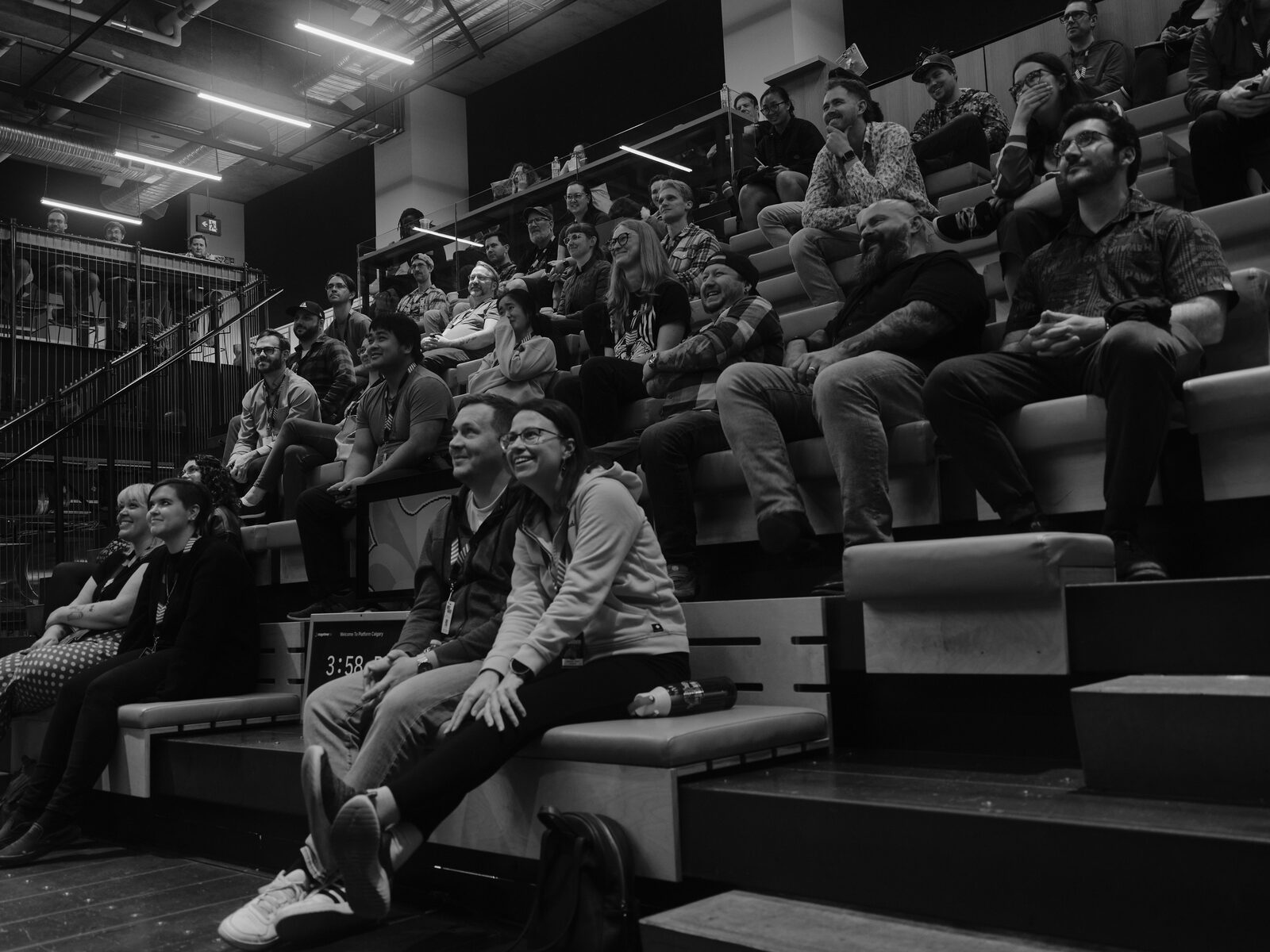Attendees on the bleachers grin and laugh during a talk, a stage monitor in front of them showing a countdown timer.