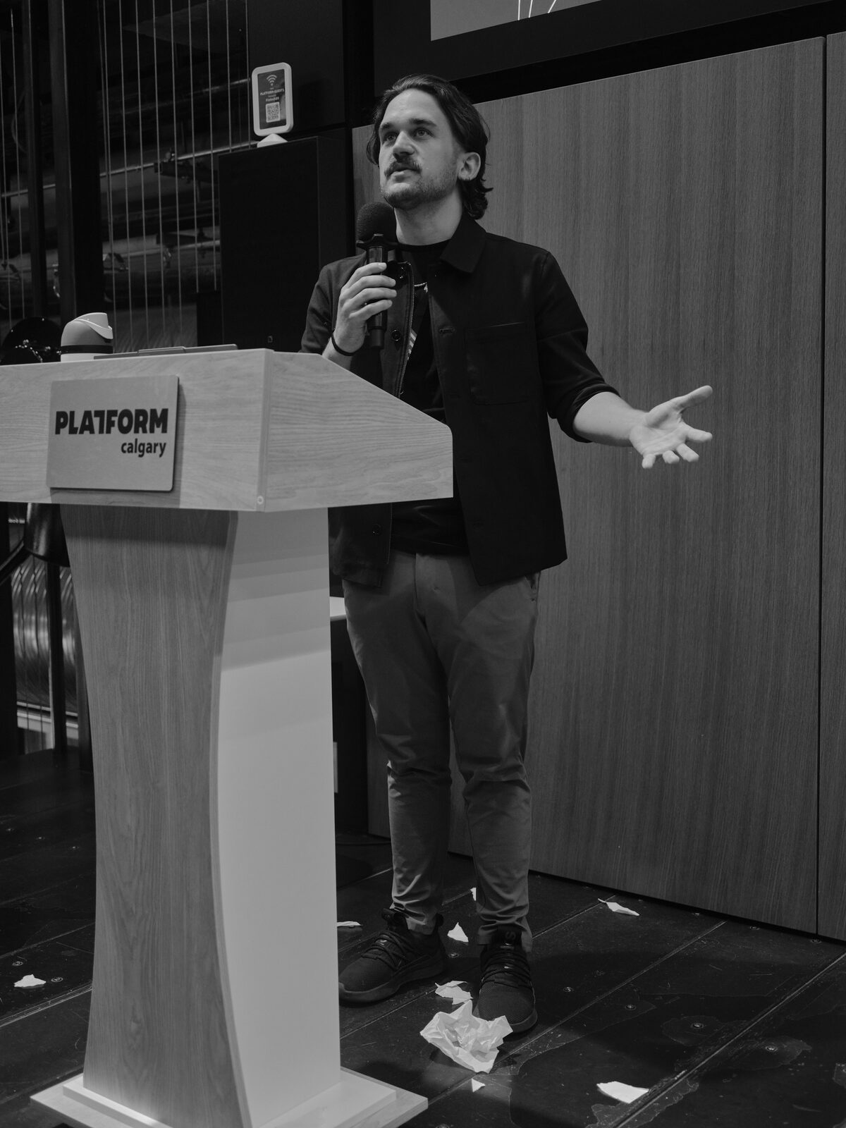 A speaker in a dark overshirt spreads one hand and looks upward beside the Platform Calgary podium, crumpled paper scattered across the stage floor.