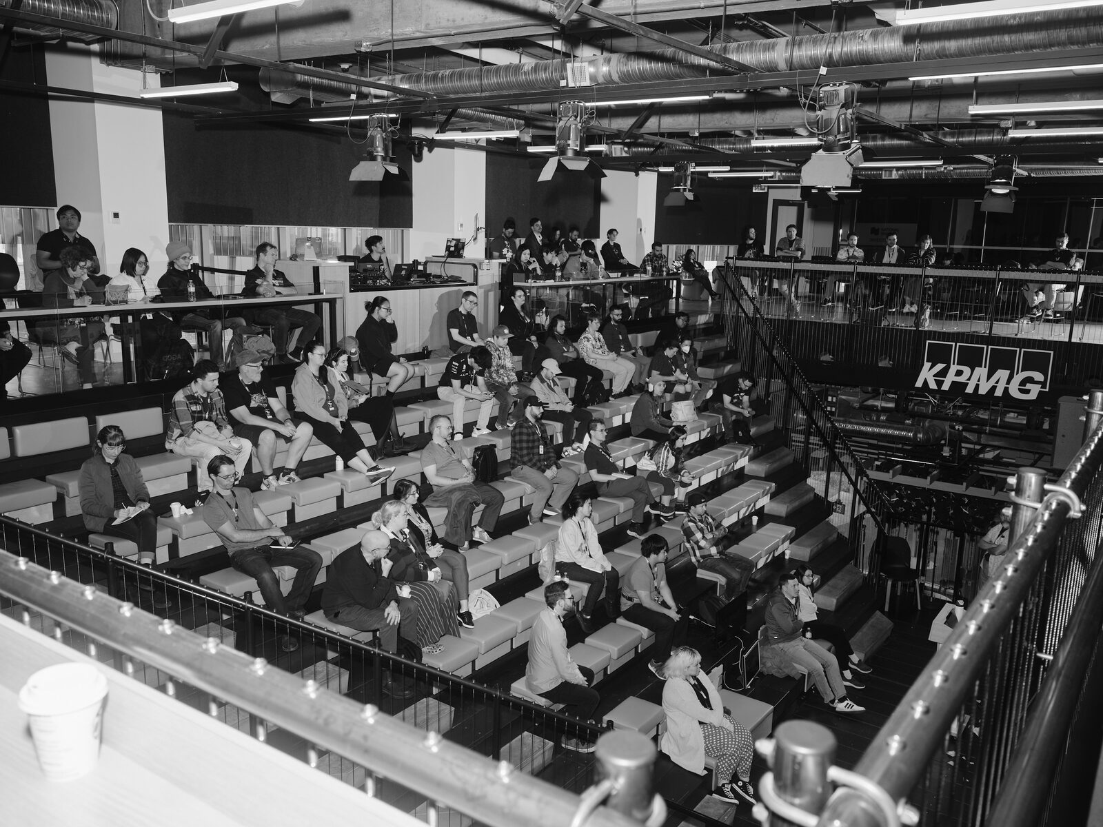 A slightly wider balcony view of the same session, the whole bleacher crowd and the upper walkway in frame, a paper cup resting on the railing in the foreground.