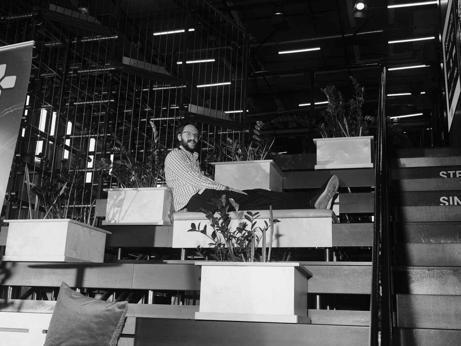 An attendee in glasses and a checked shirt stretches out on a bench among potted plants on the atrium steps, grinning at the camera.