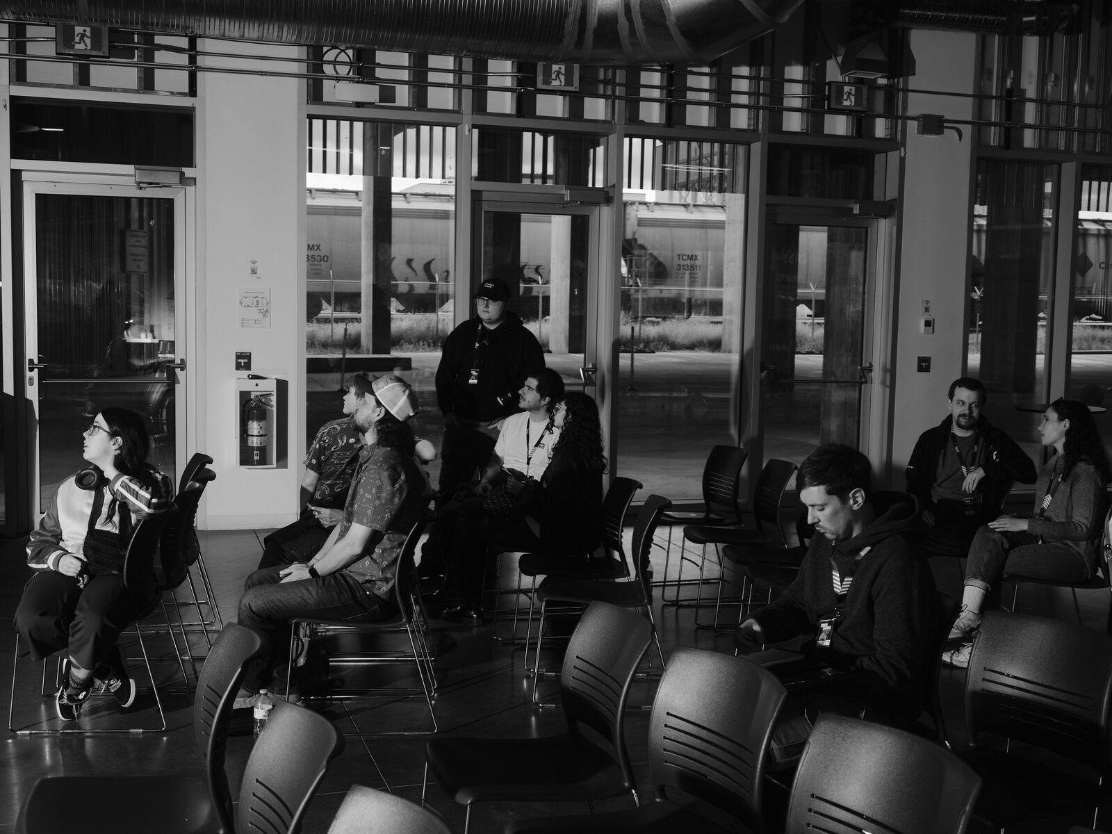 Attendees sit scattered among rows of chairs in a sunlit ground-floor room, freight train cars visible through the tall windows behind them.