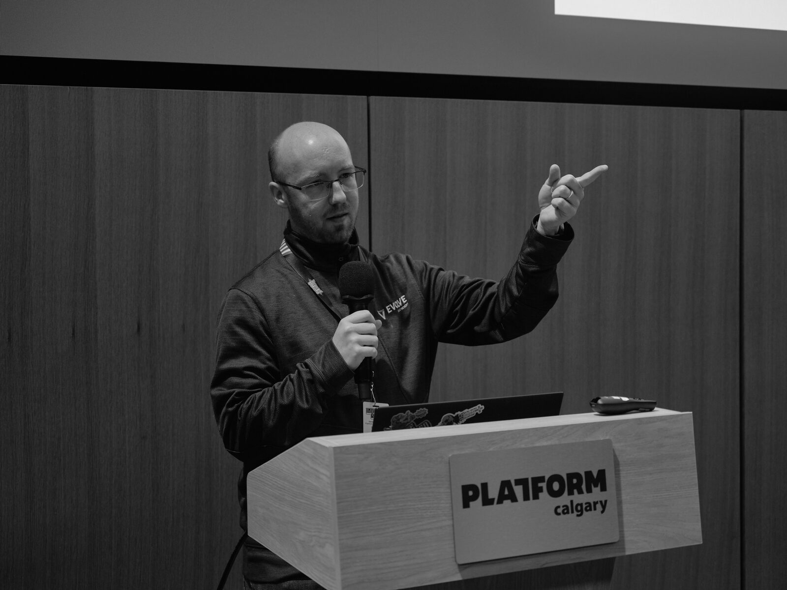 A bald speaker in glasses and a zip-up points upward with one hand, mic in the other, behind the Platform Calgary podium.