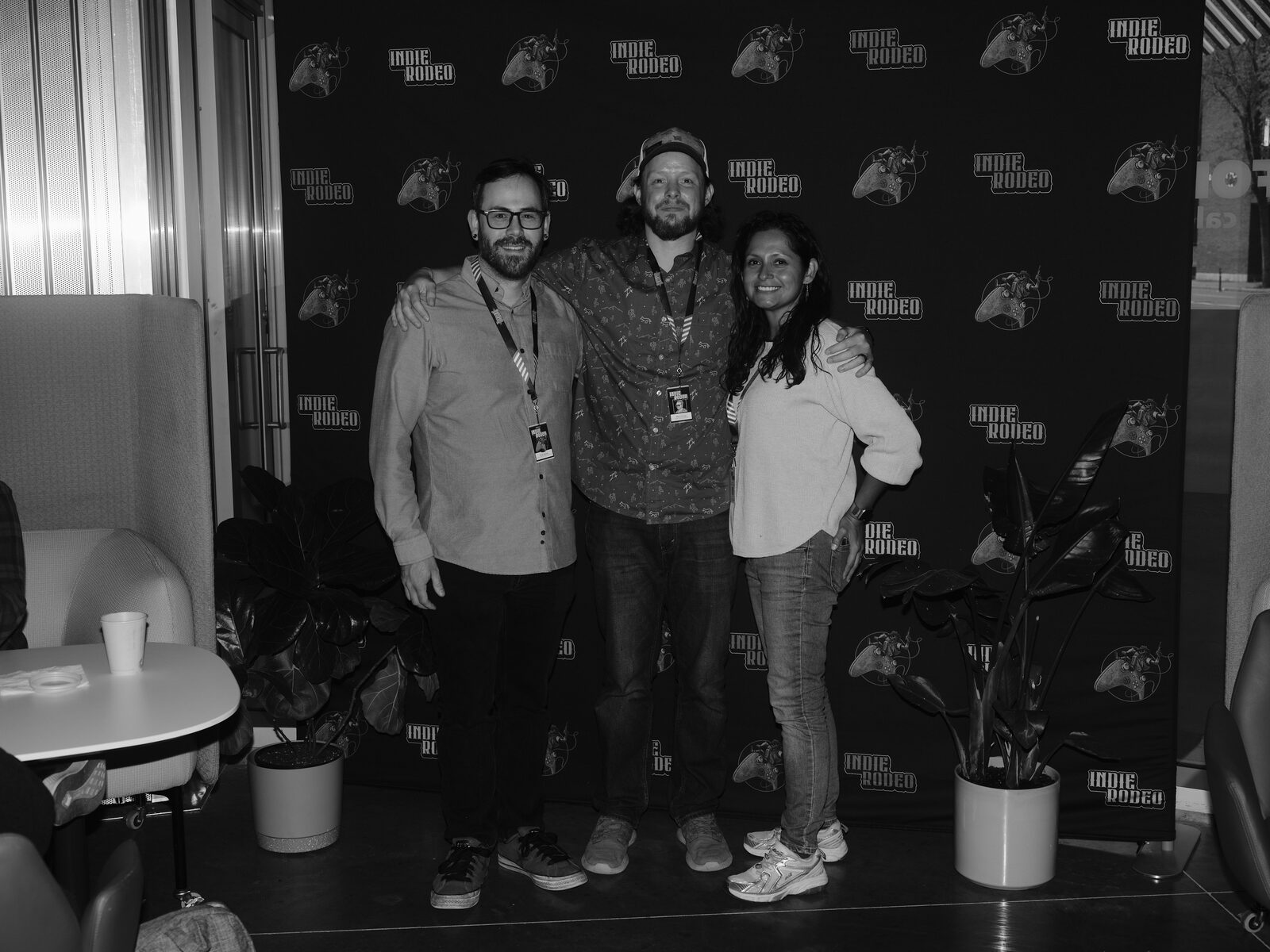 Three attendees with lanyards pose arm in arm in front of the Indie Rodeo step-and-repeat wall.
